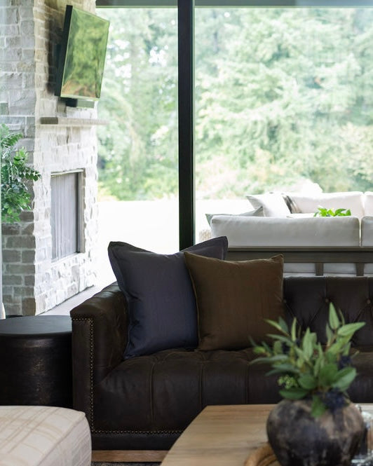 Modern living room with a black leather sofa, decorative pillows, and a view of greenery outside.