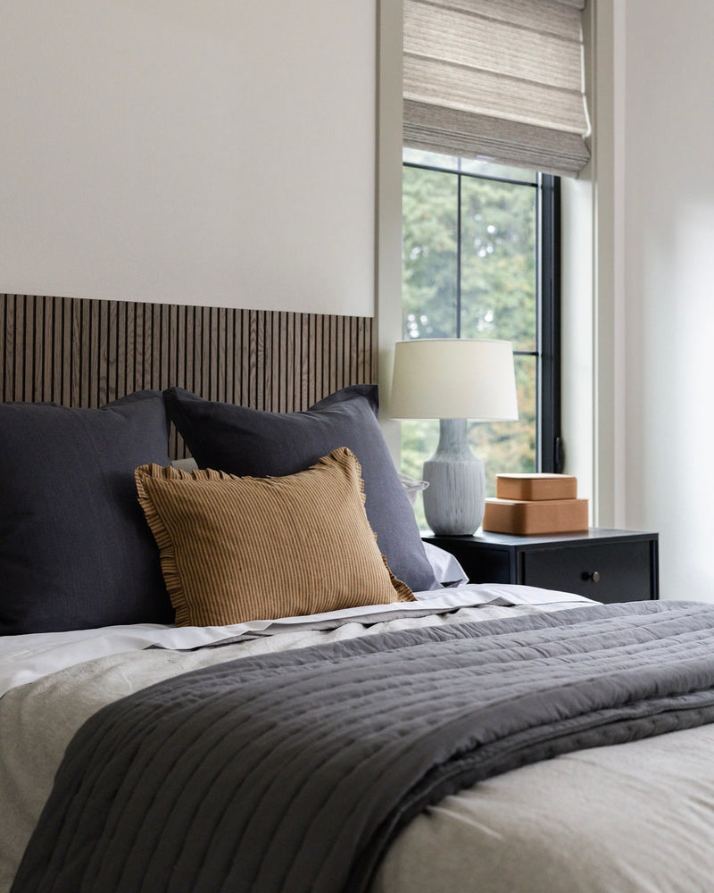 Bedroom with a bed featuring dark pillows and a gray blanket, next to a nightstand with a lamp and books.
