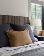 Bed with dark pillows and a brown cushion, lamp in the background